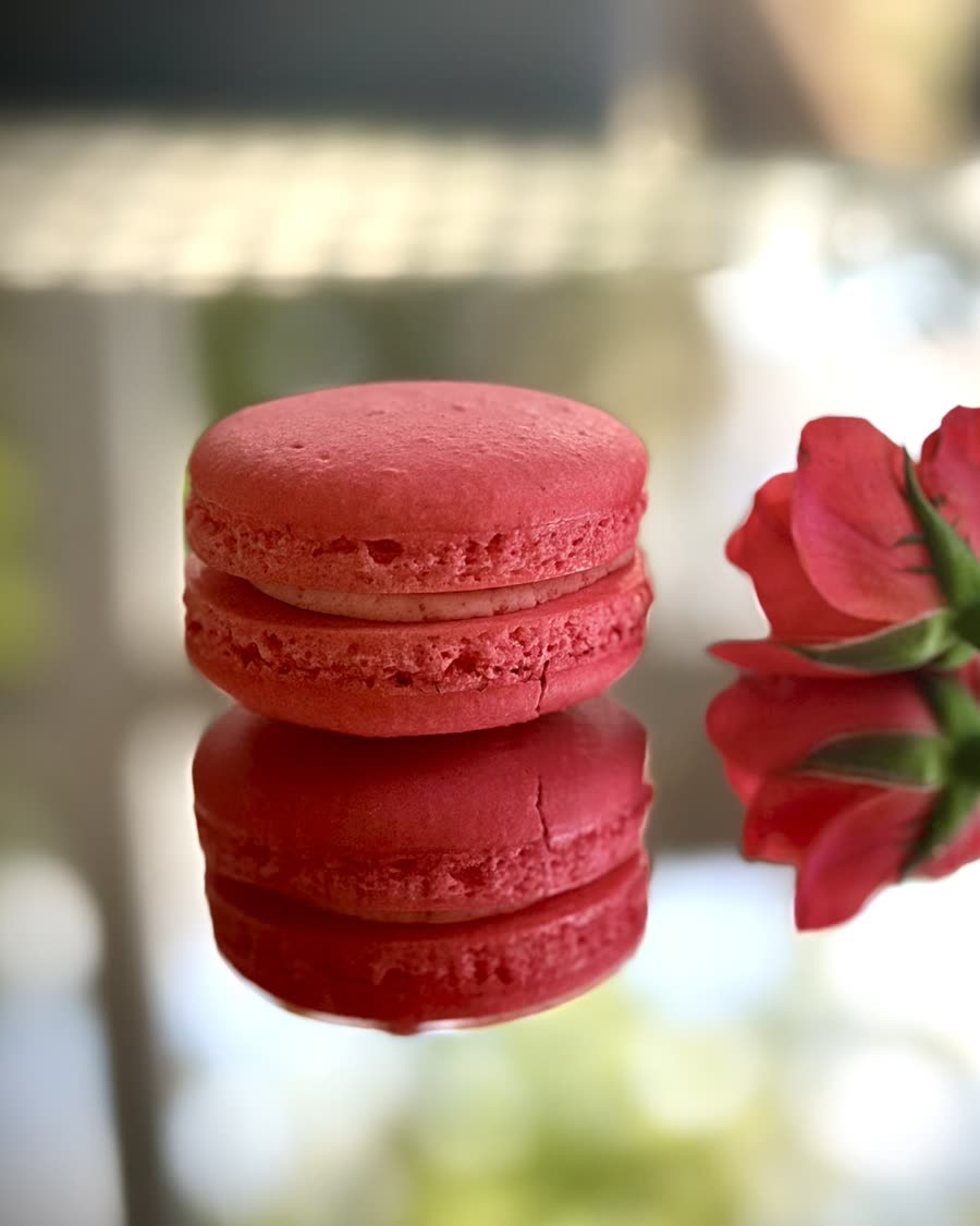 A rose-pink macaron on a mirrored table beside a red rose