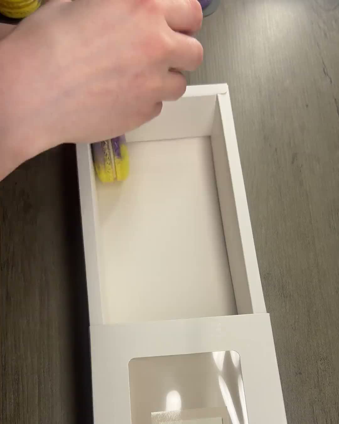 Hands placing purple and yellow macarons into a white gift box, then the finished window box full of macarons